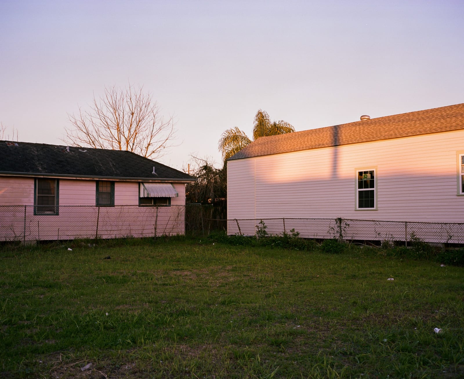 Photograph by Casey Joiner of two single story pink houses made even more pink by the setting sun. The photographer/ viewer looks across an empty and grassy backyard in the foreground at the two houses. The sky above is an ombre from a very pale blue to a medium/ light blue.