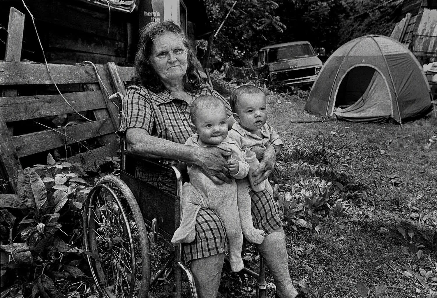 A black and white photograph by Spencer Ainsley of a woman holding two grandchildren near Sawmills, North Carolina while camping on a friend's property after losing her home in a trailer fire.