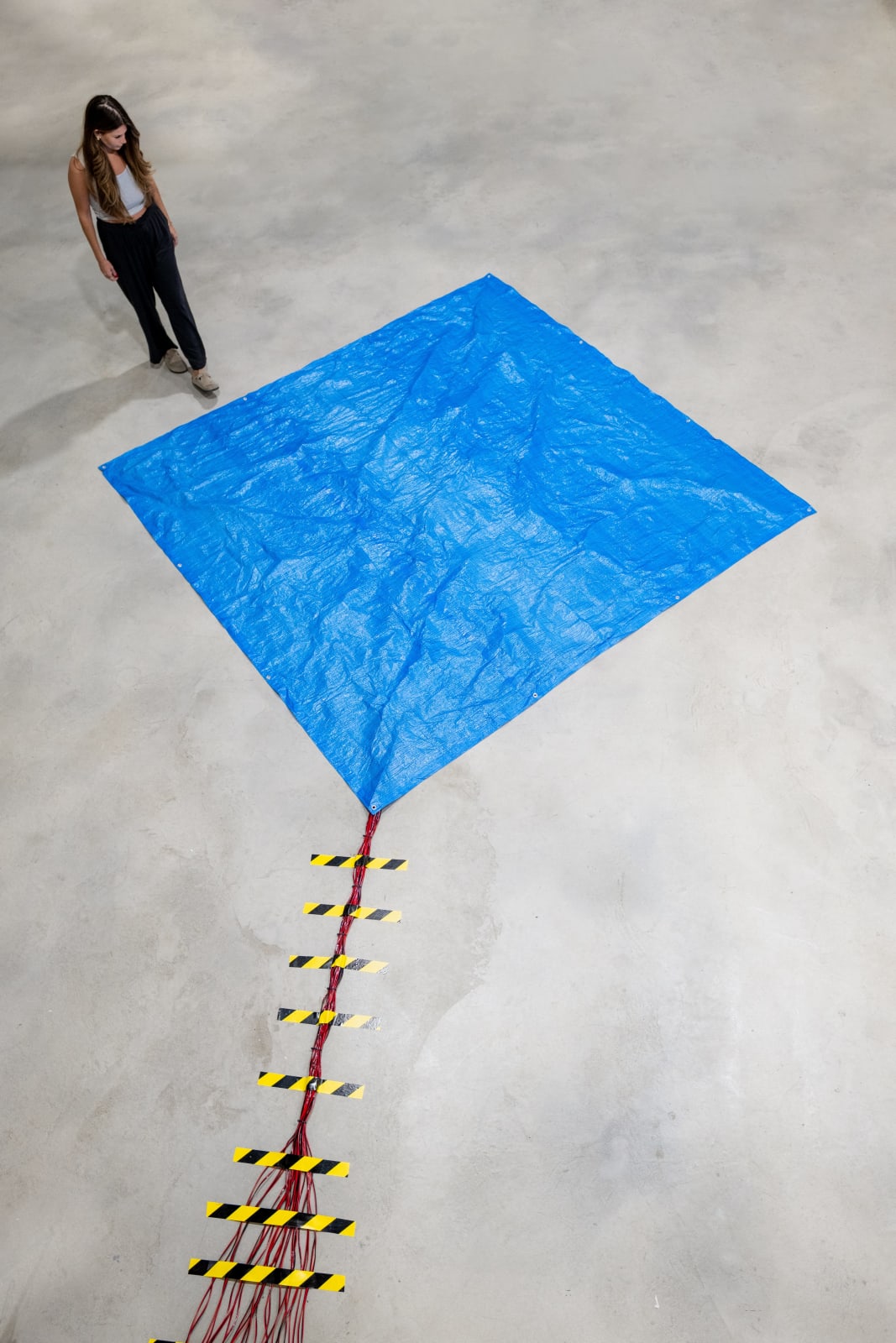 An aerial image of a sculptural installation by Ian Gouldstone, featuring a blue industrial tarp spread across a grey concrete floor. A group of red and black wires emerge from one corner of the tarp, secured to the floor by black and yellow striped tape. Not apparent from the image is the activation of the sculpture, which features 36 individual motors beneath the tarp that each randomly flick the tarp at various speeds creating a mysterious sound at randomized tempos. The sound is reminiscent of water drops falling from a leaky roof.