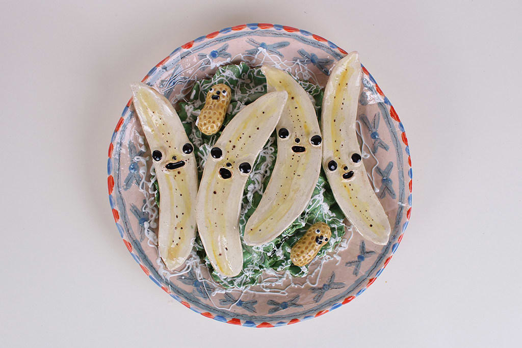 Ceramic sculpture of a bowl of bananas cut in half with faces and two peanuts with faces on a bed of lettuce