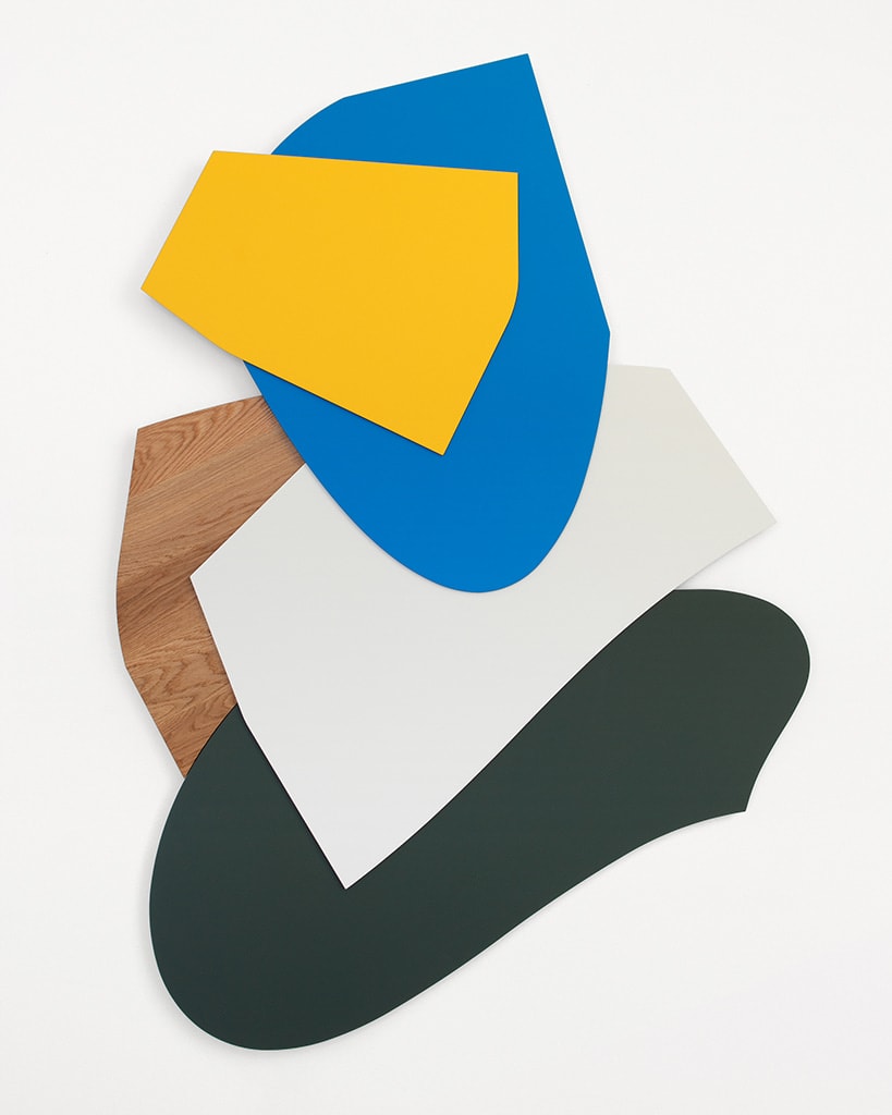 An abstract wall hanging sculpture with irregular shapes stacked on top of each other. There is a dark green oval shape on the bottom, a white square shape layered on top of that, a blank wooden piece is tucked behind the white and green piece. On top of the white piece is an irregular circle blue shape and on top of that is a bright yellow square piece.