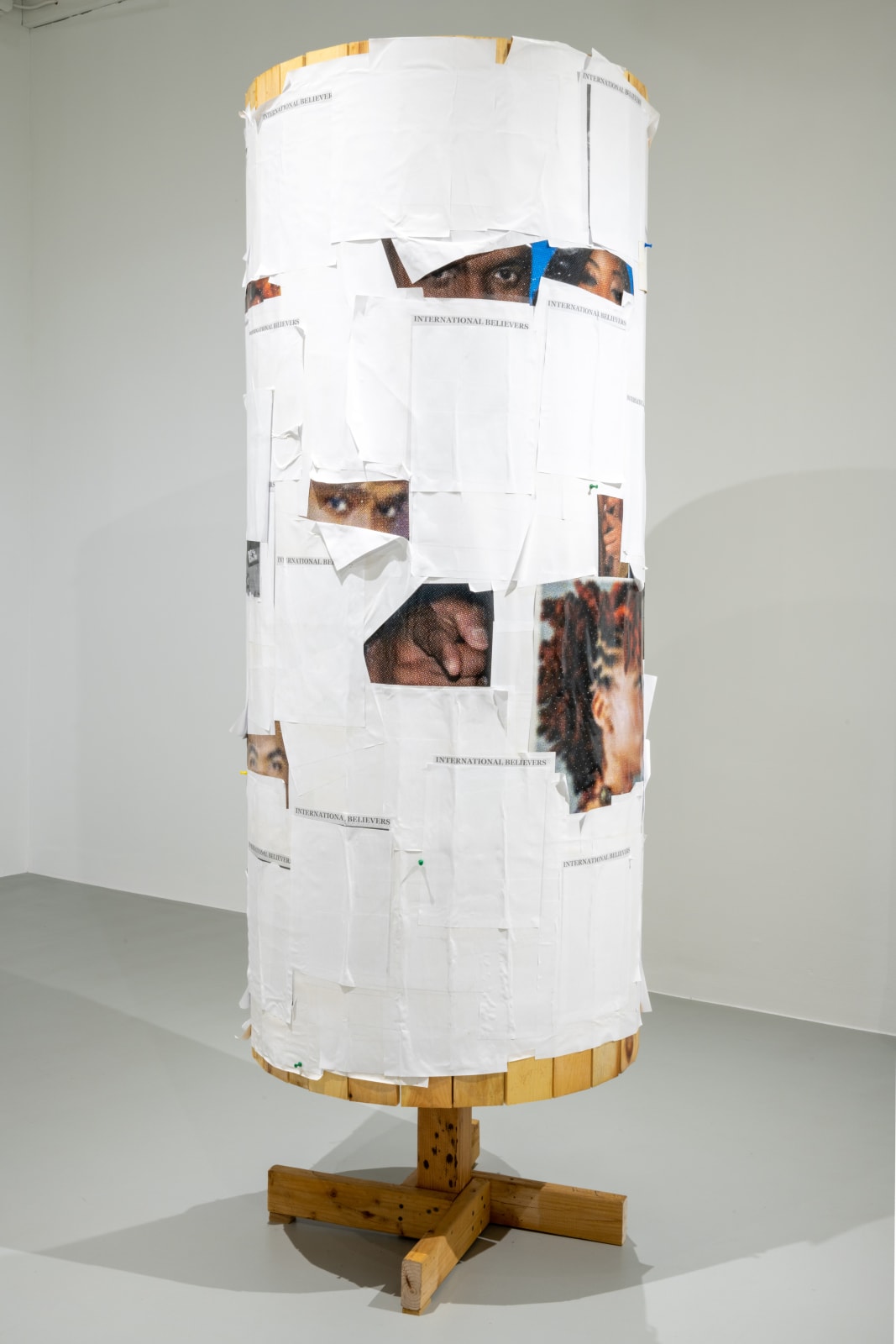 Round freestanding wood sculpture with multiple close up images of human figures adhered to the surface and obscured by pieces of white paper