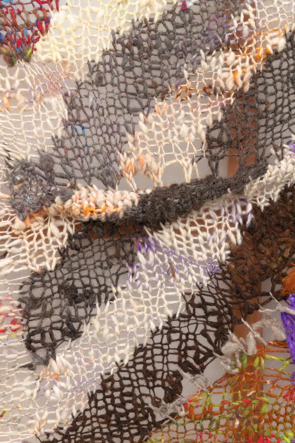 A close-up detail of geometric composition in multi-colored fibers stretched across a redwood frame as one would stretch a painting. The fibers are knit at varying thicknesses so that in places one can see through the stitches to the wall behind