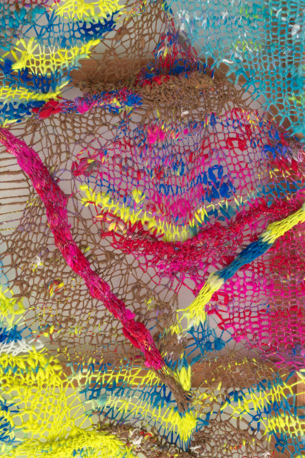 A detail of an organic composition in multi-colored fibers stretched across a redwood frame as one would stretch a painting. The fibers are knit at varying thicknesses so that in places one can see through the stitches to the wall behind.