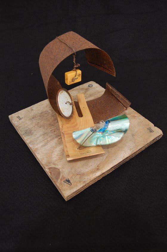 Assemblage sculpture on wooden base with a broken CD and a yellow block hanging from a brown curve