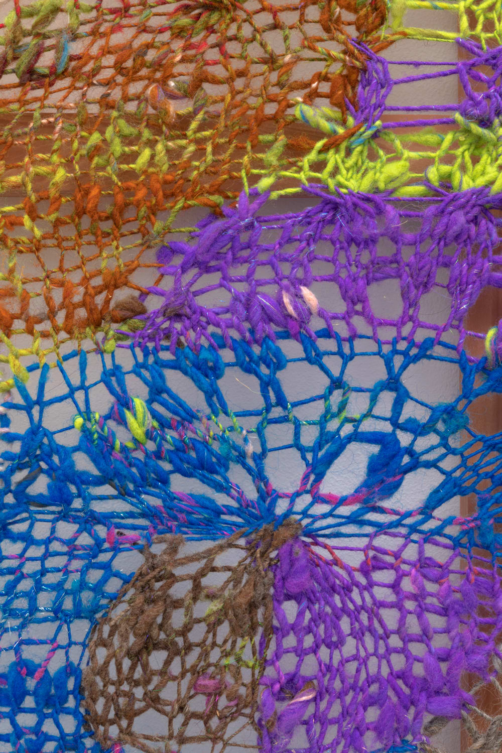 A close-up detail of geometric composition in multi-colored fibers stretched across a redwood frame as one would stretch a painting. The fibers are knit at varying thicknesses so that in places one can see through the stitches to the wall behind
