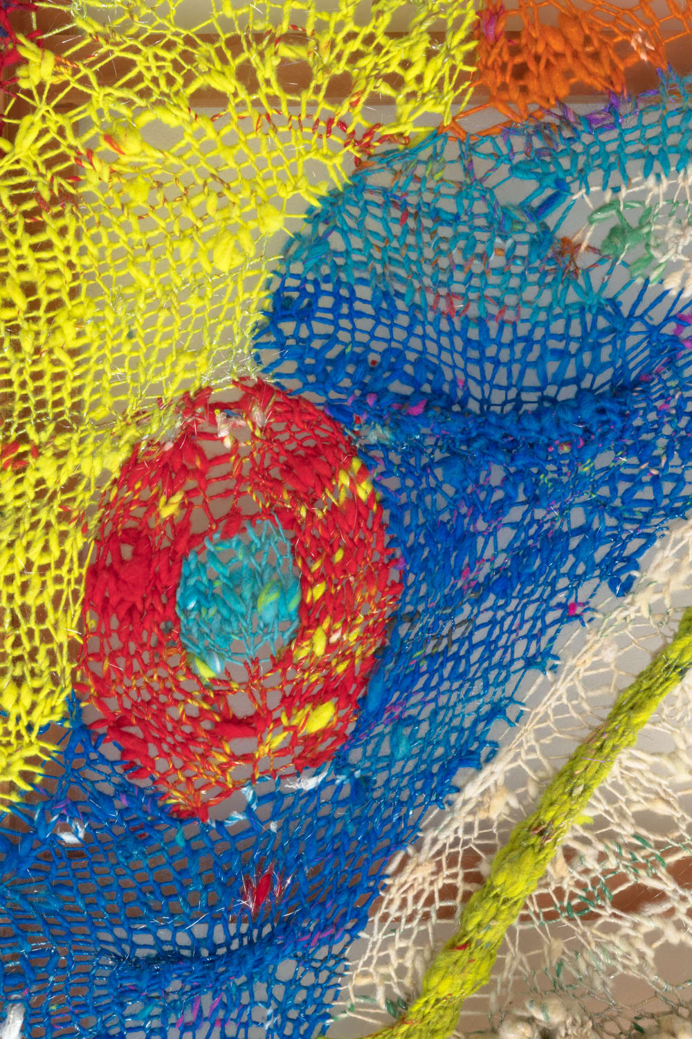 A detail of an organic composition in multi-colored fibers stretched across a redwood frame as one would stretch a painting. The fibers are knit at varying thicknesses so that in places one can see through the stitches to the wall behind.
