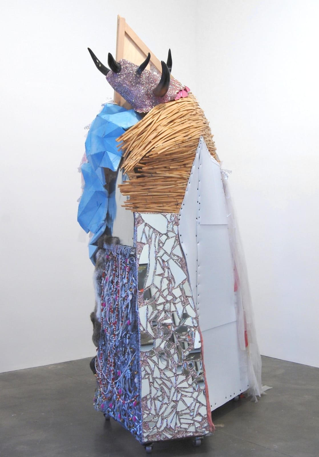 Vertical sculpture sitting on the floor comprised of wood, screws, chicken wire, casters, plastic, silicone, foam,plaster, coroplast, mirrors, glitter, crystals, aluminum, copper, brass, bamboo, nKisi head figure, Nok beads, polish beads, clothes, fabric trim, oil paint, ink on paper, rabbit fur, elephant tusk, bull horns