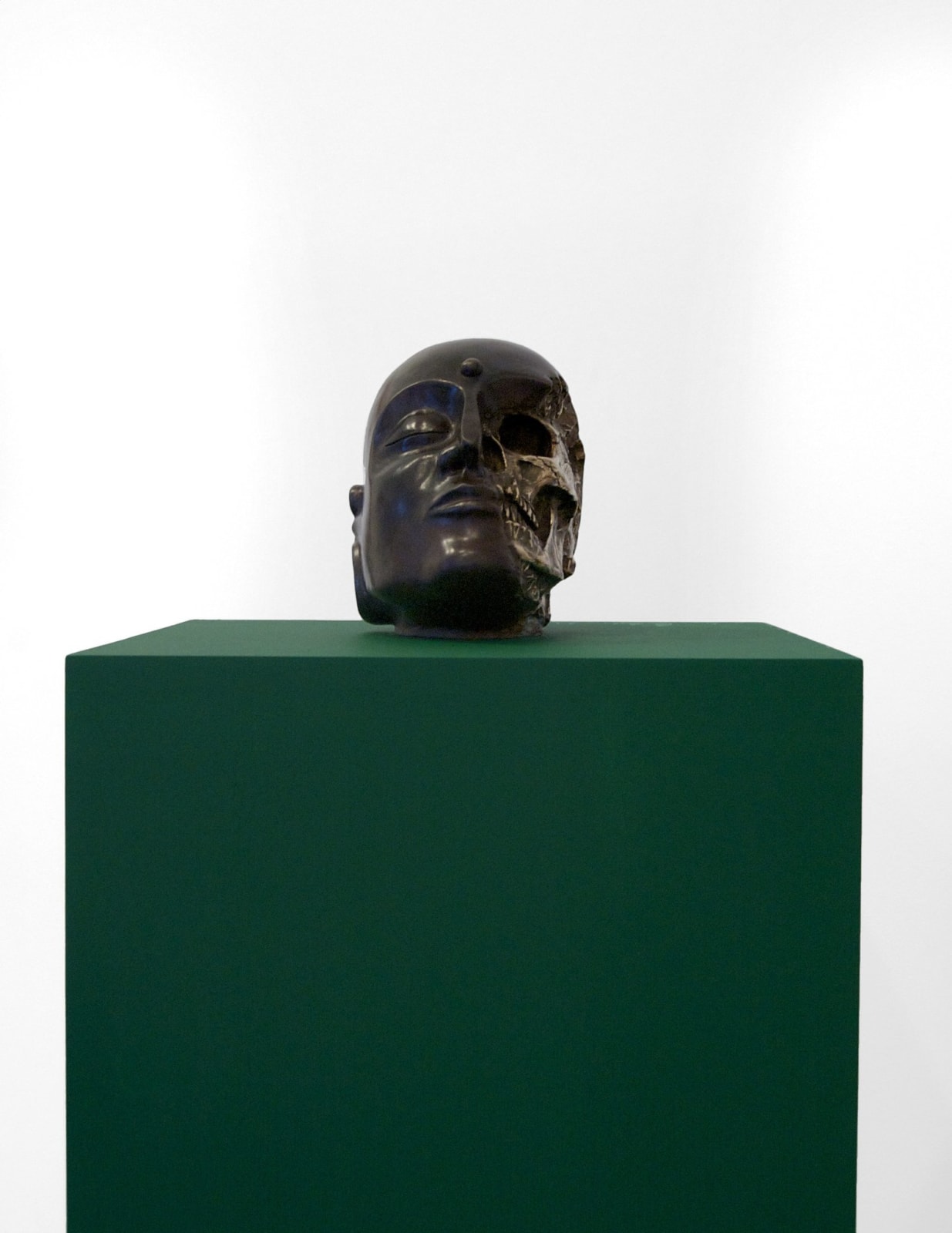 Bronze sculpture of half head, half skeleton, sitting on top of a dark green platform