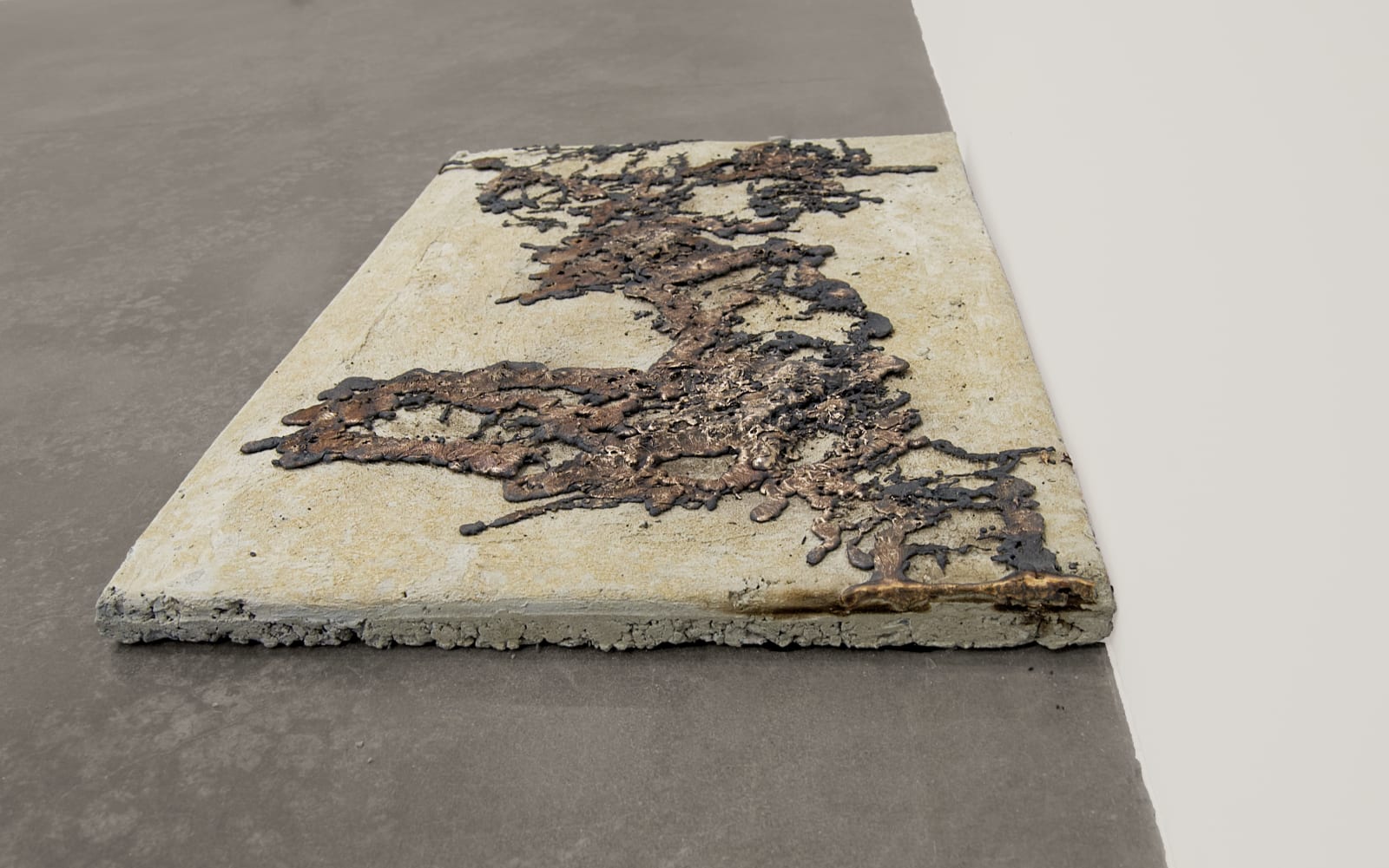 A concrete and bronze sculpture in the size and shape of a sidewalk square sits in a gallery on a concrete floor.