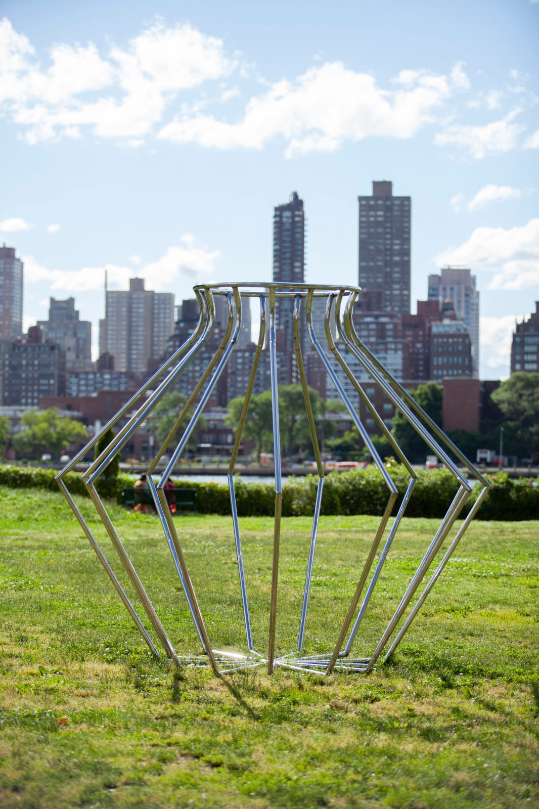 Outdoor installation view of a metal sculpture that resembles an angular vessel set against a city skyline at Socrates Sculpture Park