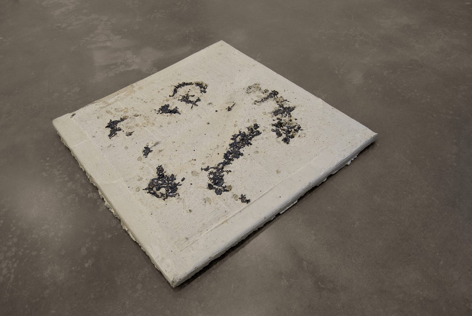 A close up of two bronze and concrete sculptures resting on top of one another.A concrete and bronz sculpture in the size and shape of a sidewalk square sits in a gallery on a concrete floor