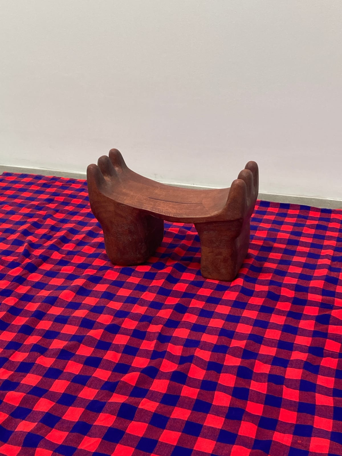 A wooden headrest with thick, stout legs and three finger-like rounded divisions curving up on either side on a red and blue patterned cloth