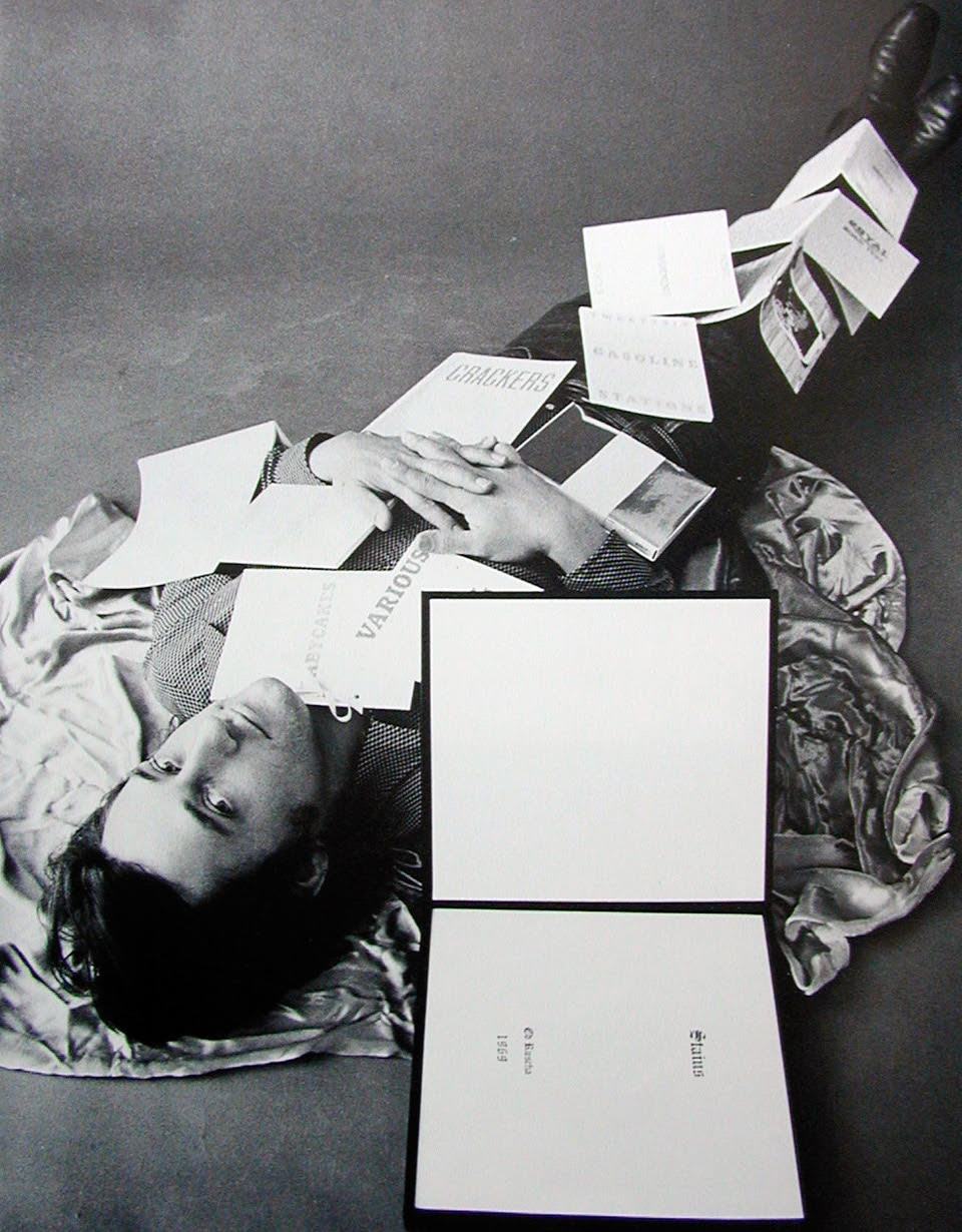 Black and white image of dark haired man lying underneath pile of books