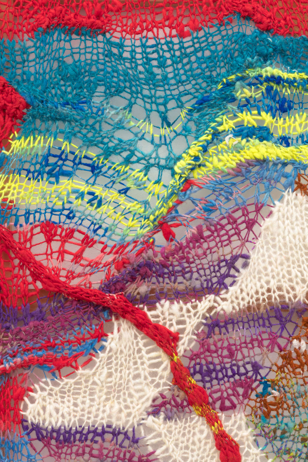 A detail of a cacophonous composition in multi-colored fibers stretched across a redwood frame as one would stretch a painting. The fibers are knit at varying thicknesses so that in places one can see through the stitches to the wall behind. The composition has a wave-like pattern with many fractal fields of varying colors