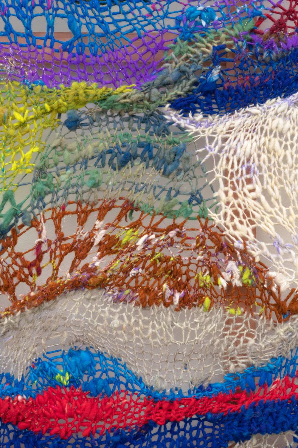 A close-up detail of a cacophonous composition in multi-colored fibers stretched across a redwood frame as one would stretch a painting. The fibers are knit at varying thicknesses so that in places one can see through the stitches to the wall behind. The composition has a wave-like pattern with many fractal fields of varying colors