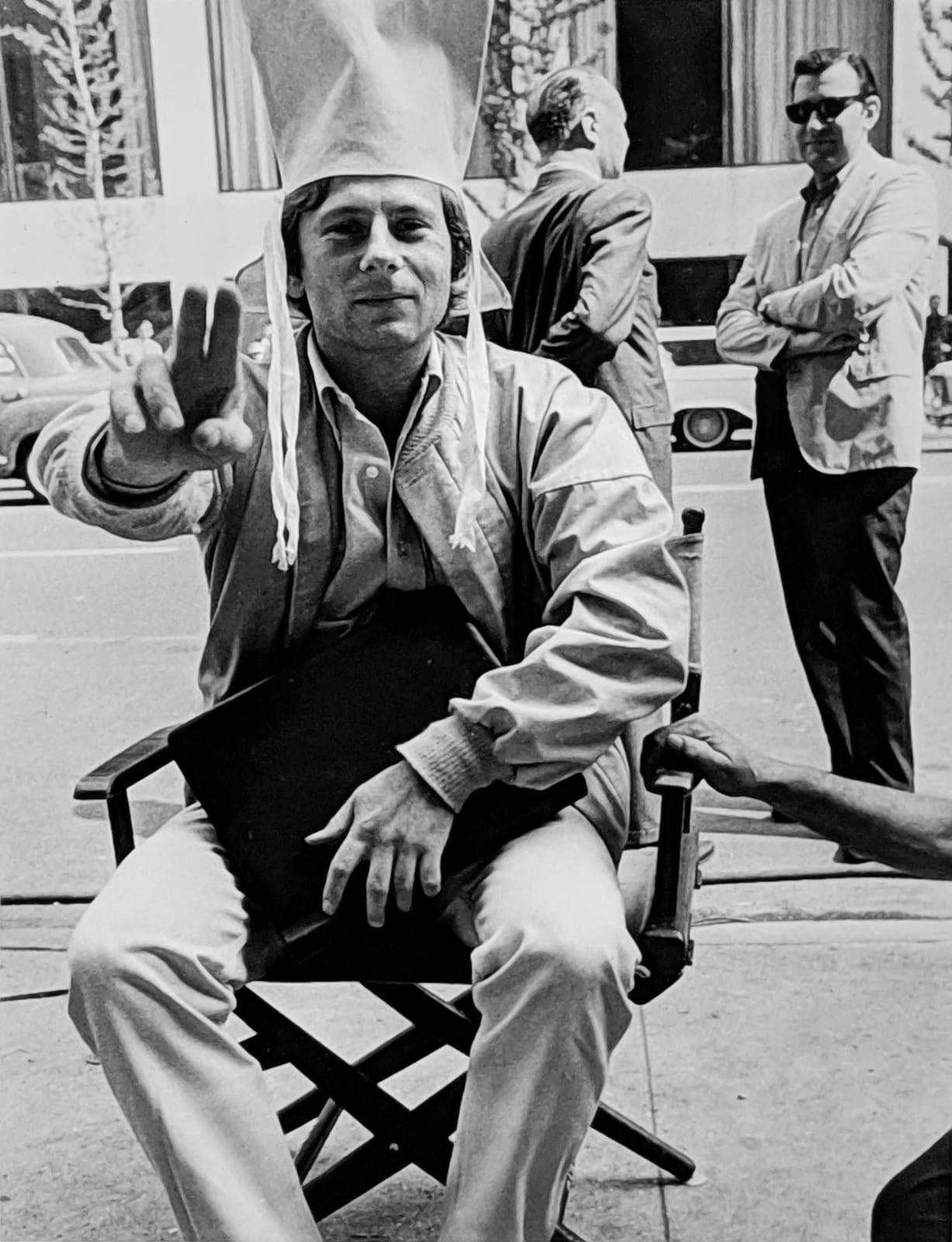 Black and white photograph of Roman Polanski taking a break between takes,holding two fingers to the camera and smirking, on the set of “Rosemary's Baby”