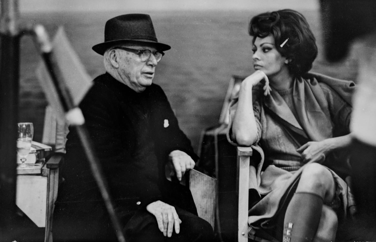 Black and white photograph of Sophia Loren sitting and wearing a dressing robe, looking at Charlie Chaplin on the set of “A Countess from Hong Kong”