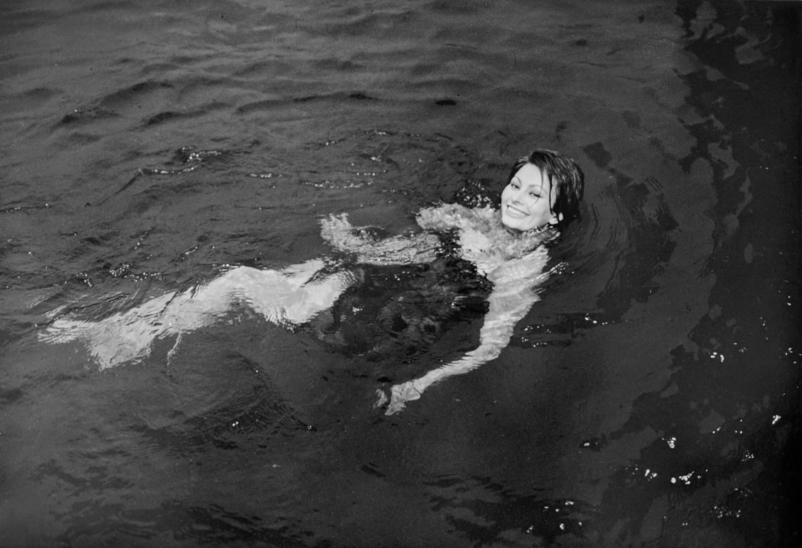 Black and white photograph of Sophia Loren swimming and looking up to smile at the camera, relaxing between takes on the set of “A Countess from Hong Kong”