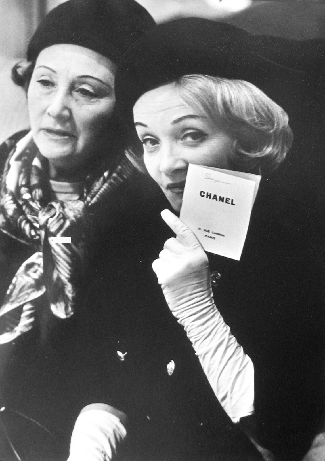 Black and white photograph of Marlene Dietrich sitting next to a patron holding a Chanel card at a fashion presentation at the House of Chanel