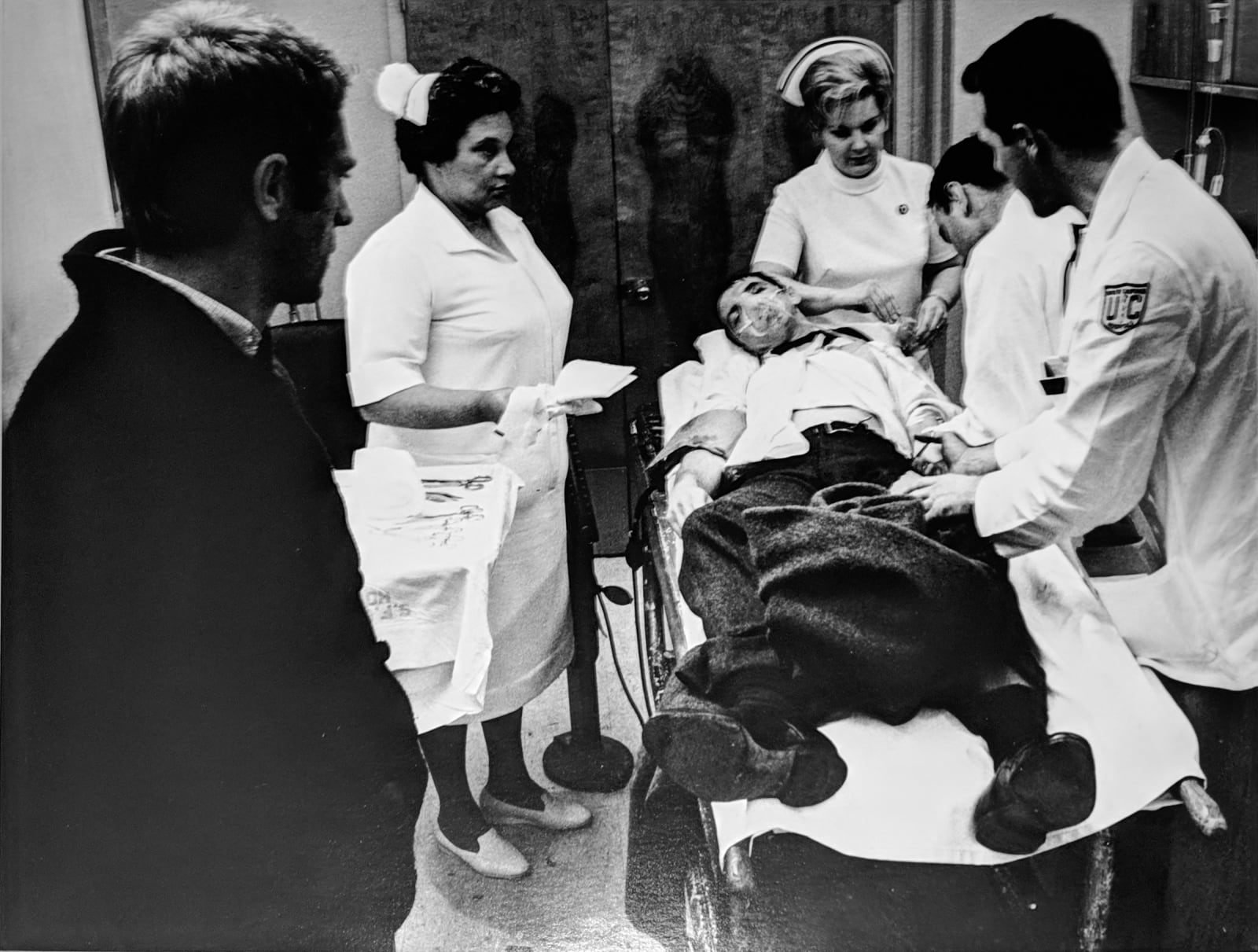 Black and white photograph of Steve Mcqueen talking with actors dressed as nurses and doctors, with an extra in a stretcher, in between takes on the set of “Bullitt”