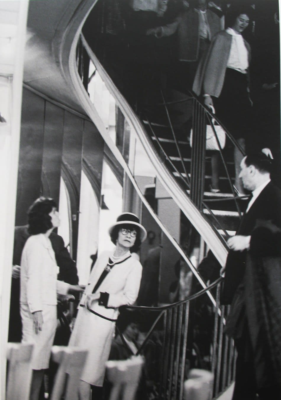 Black and white photograph of Designer Coco Chanel leaning against the staircase in the House of Chanel in Paris