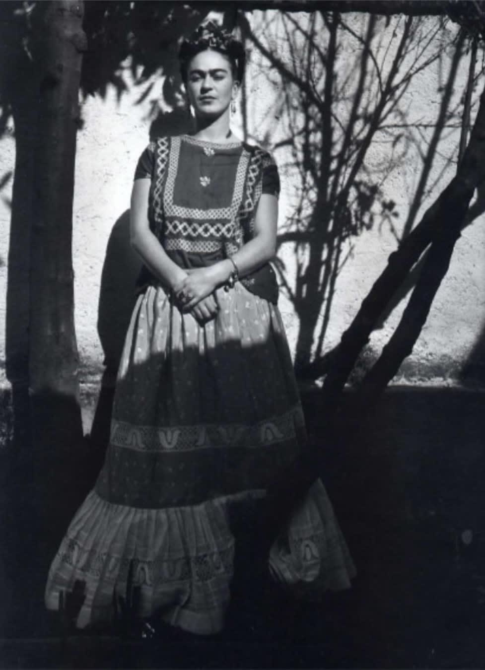 Black and white photograph of artist Frida Kahlo in front of an exterior wall of her home and studio, the Blue House, in between shadows cast by trees