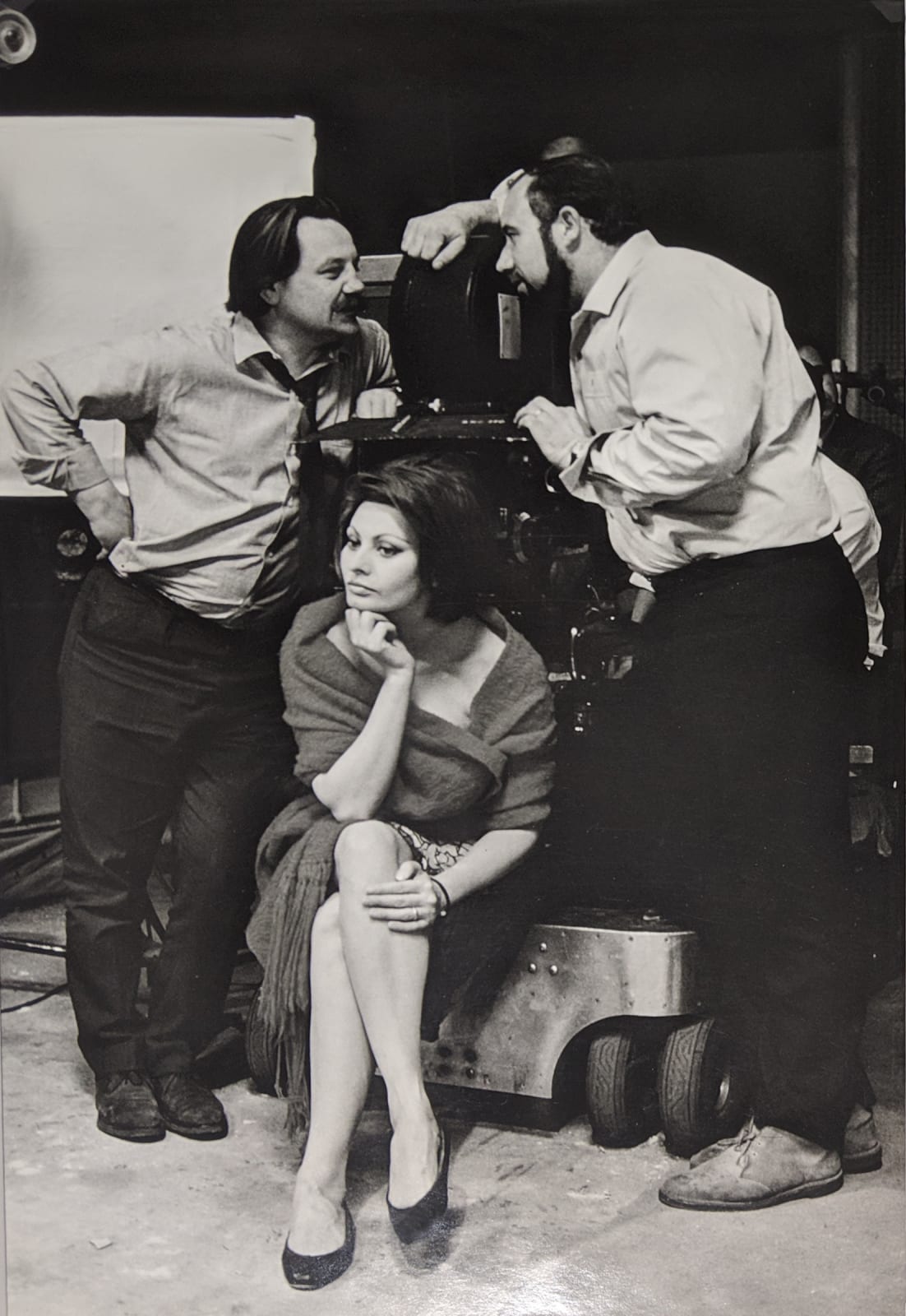 Black and white photograph of Sofia Loren sitting down with two crew members standing over her and speaking to each other, on the set of “A Countess from Hong Kong”