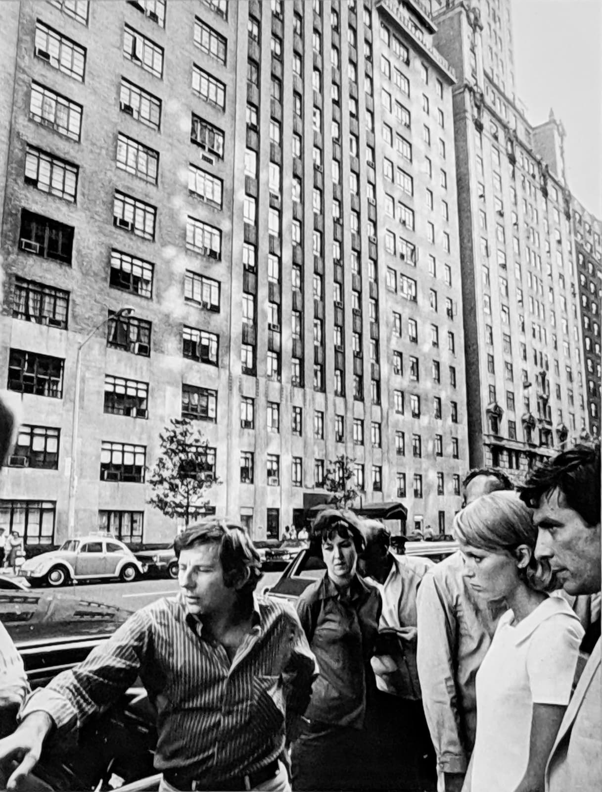 Black and white photograph of Roman Polanski, Mia Farrow, John Cassavetes and crew in front of The Dakota building,with large apartment buildings in the background in New York City, on the set of “Rosemary's Baby”