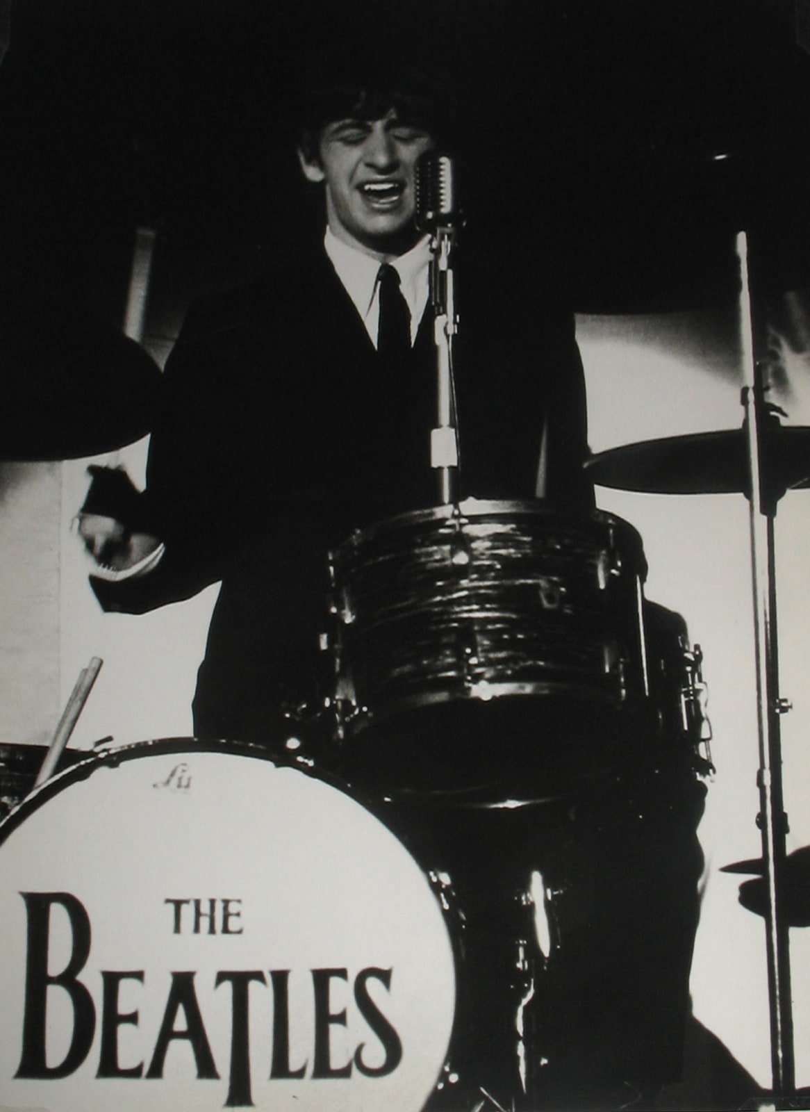 Black and white photograph of Ringo Starr playing the drums, the bass drum has “The Beatles” written on it.