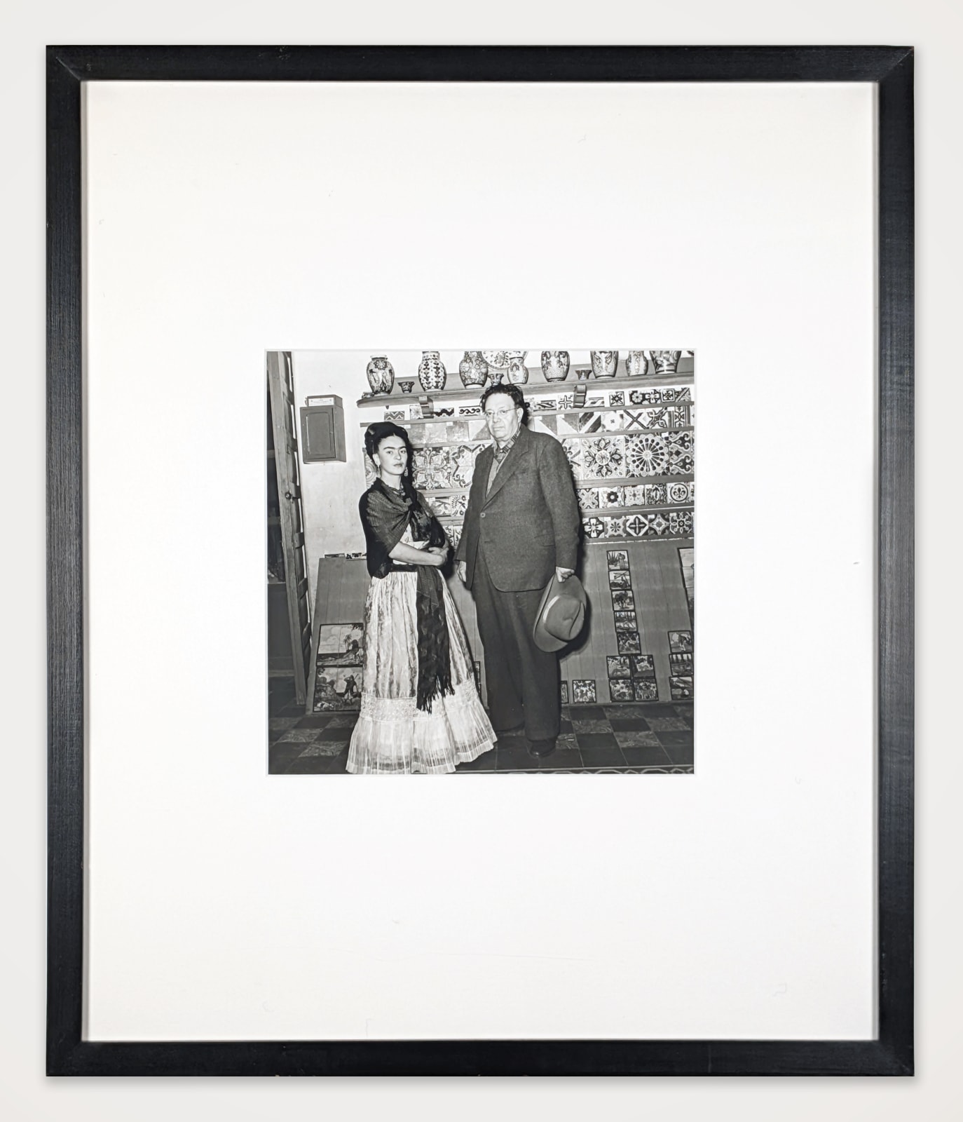 Framed black and white photograph of Frida Kahlo and Diego Rivera posing in front of a tile-covered wall inside the Blue House