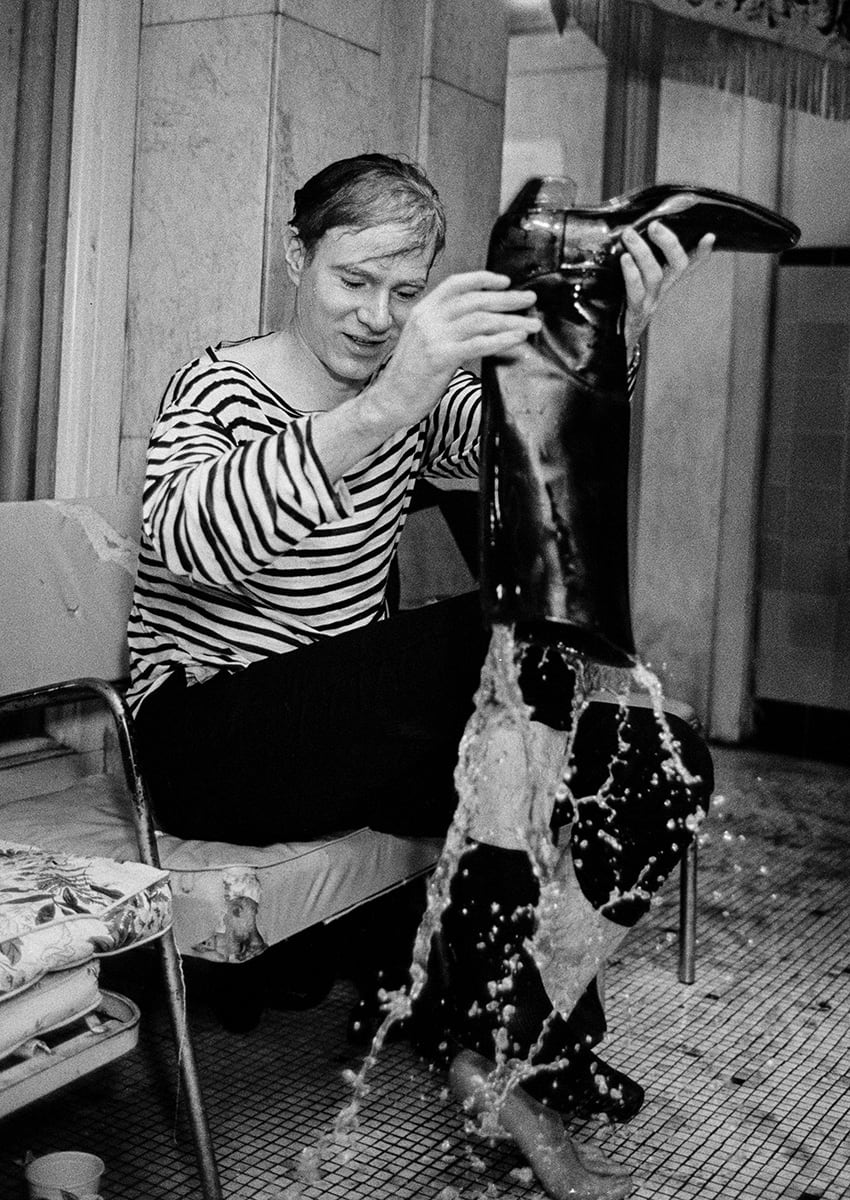Black and white photograph of Andy Warhol soaked and emptying his boot full of water onto the ground
