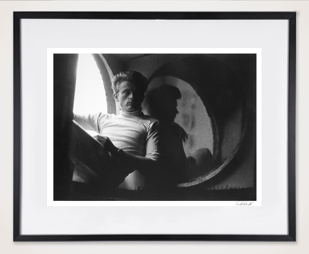 Framed black and white photograph of James Dean sitting by a window holding a book in an apartment setting