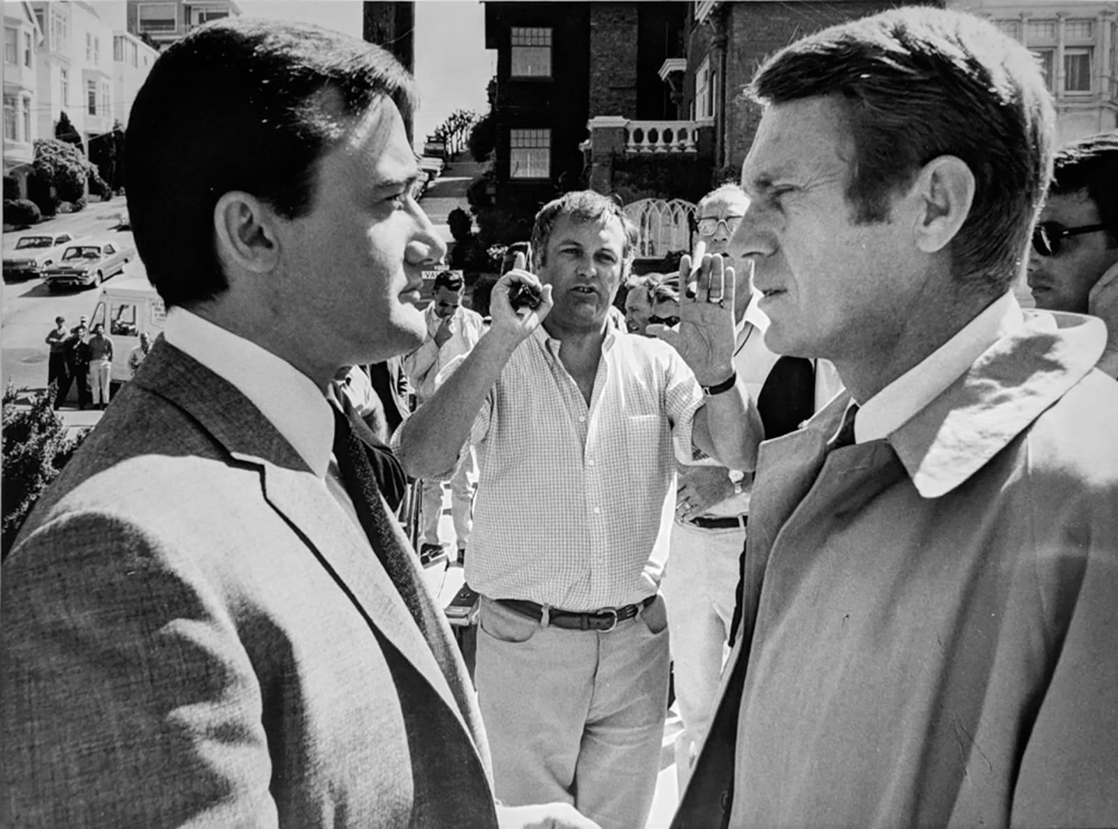 Black and white photograph of Robert Vaughn and Steve McQueen facing each other in profile in the foreground, and director Peter Yates in the background on the phone, setting up a shot, on the set of “Bullitt”