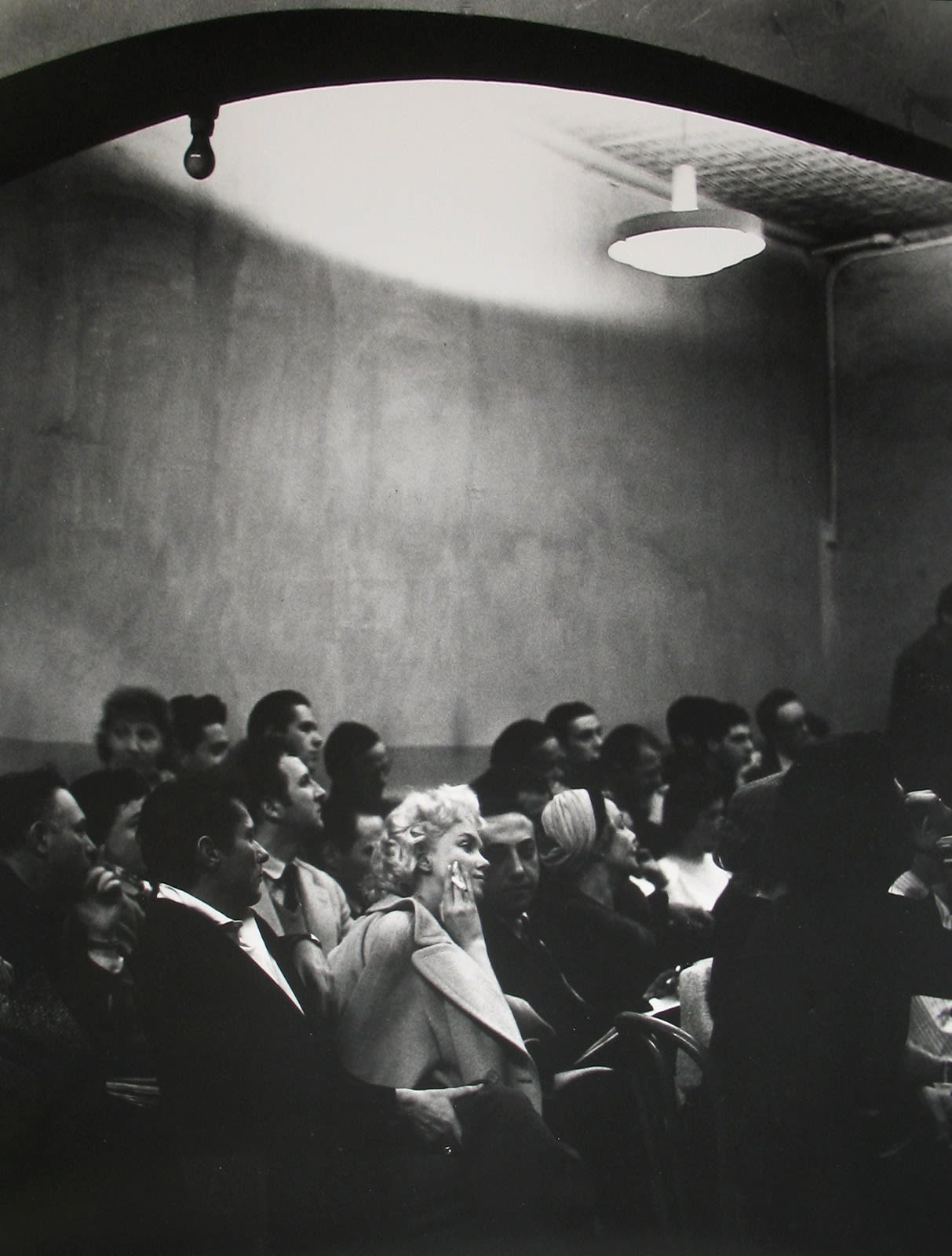 Black and white photograph of Marilyn Monroe holding a cigarette in a light-colored coat sitting amongst a crowd of other figures dressed in dark colors within a studio setting
