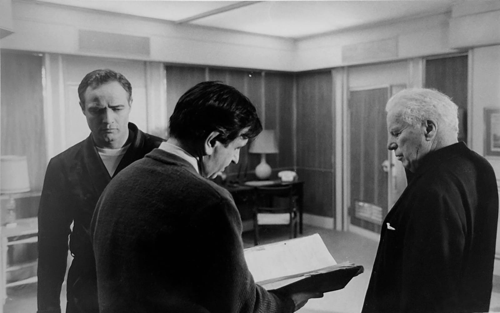 Black and white photograph of Charlie Chaplin and Marlon Brando and a man holding a script, discussing a scene on the set of “A Countess from Hong Kong”