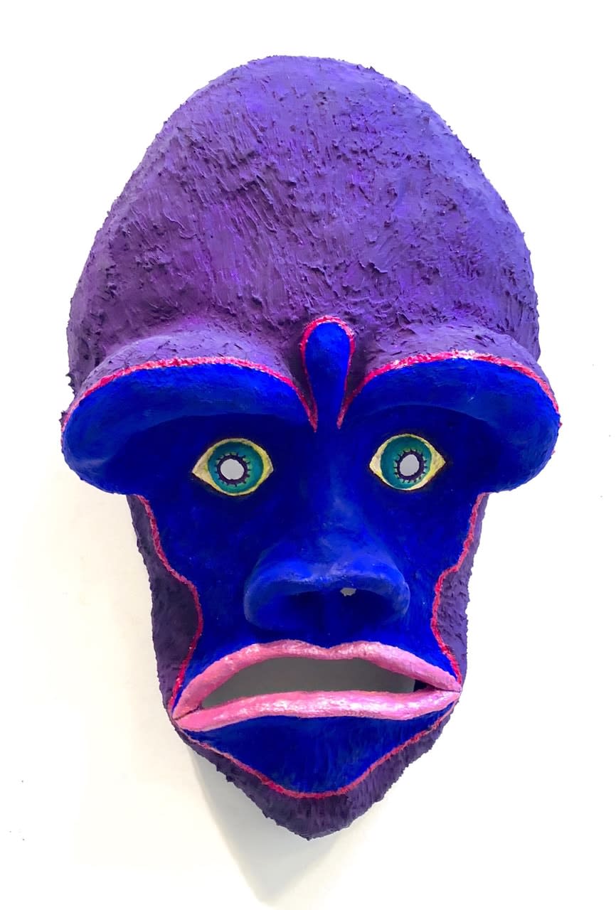 A vibrant violet mask made of acyrlic on plaster is mounted on a wall and bears humanoid features including large green eyes, and pink lips.
