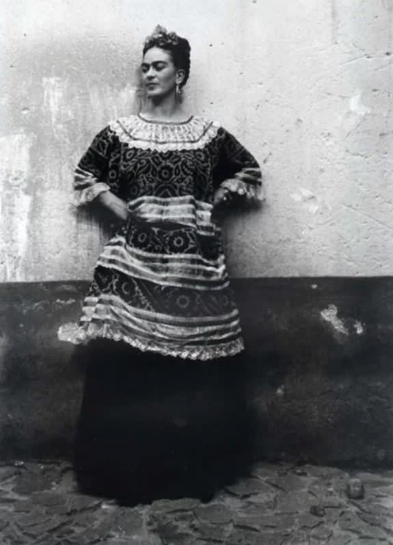 Black and white photograph of Frida Kahlo in traditional Tehuana dress leaning against a bicolored wall with her hands on her hips outside the Blue House in Mexico City
