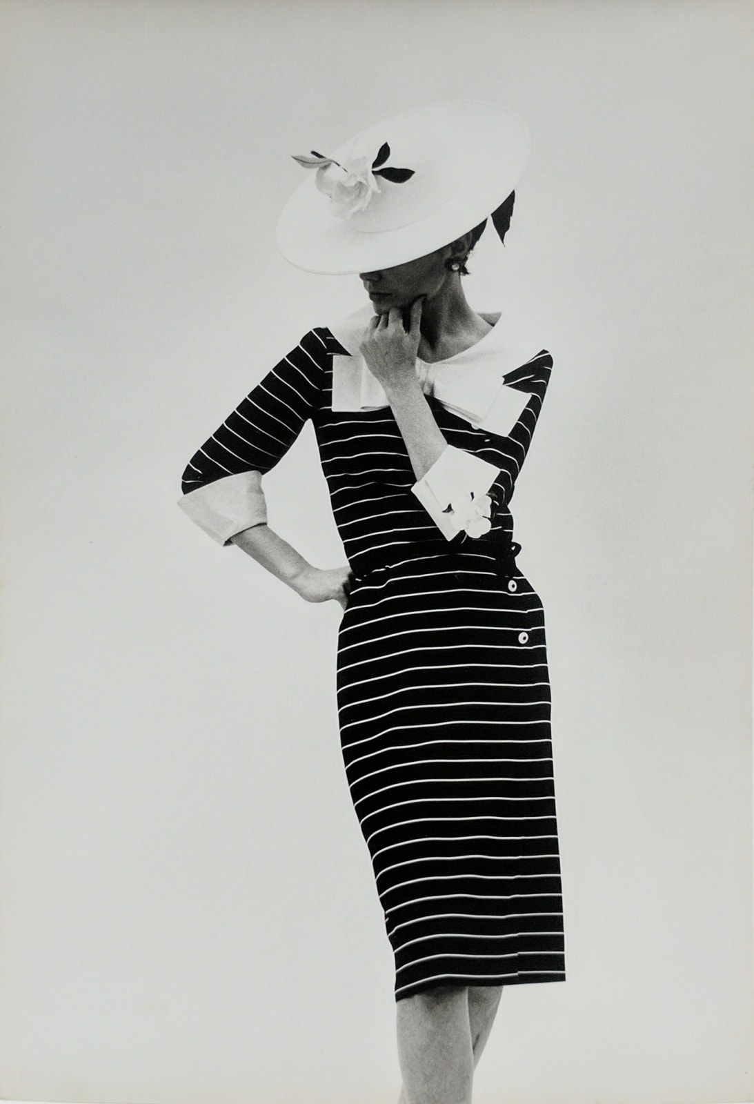 Black and white photograph of a Chanel model in a studio posing in a black and white chanel dress and a large white hat, looking down and to the left so that the hat covers her face