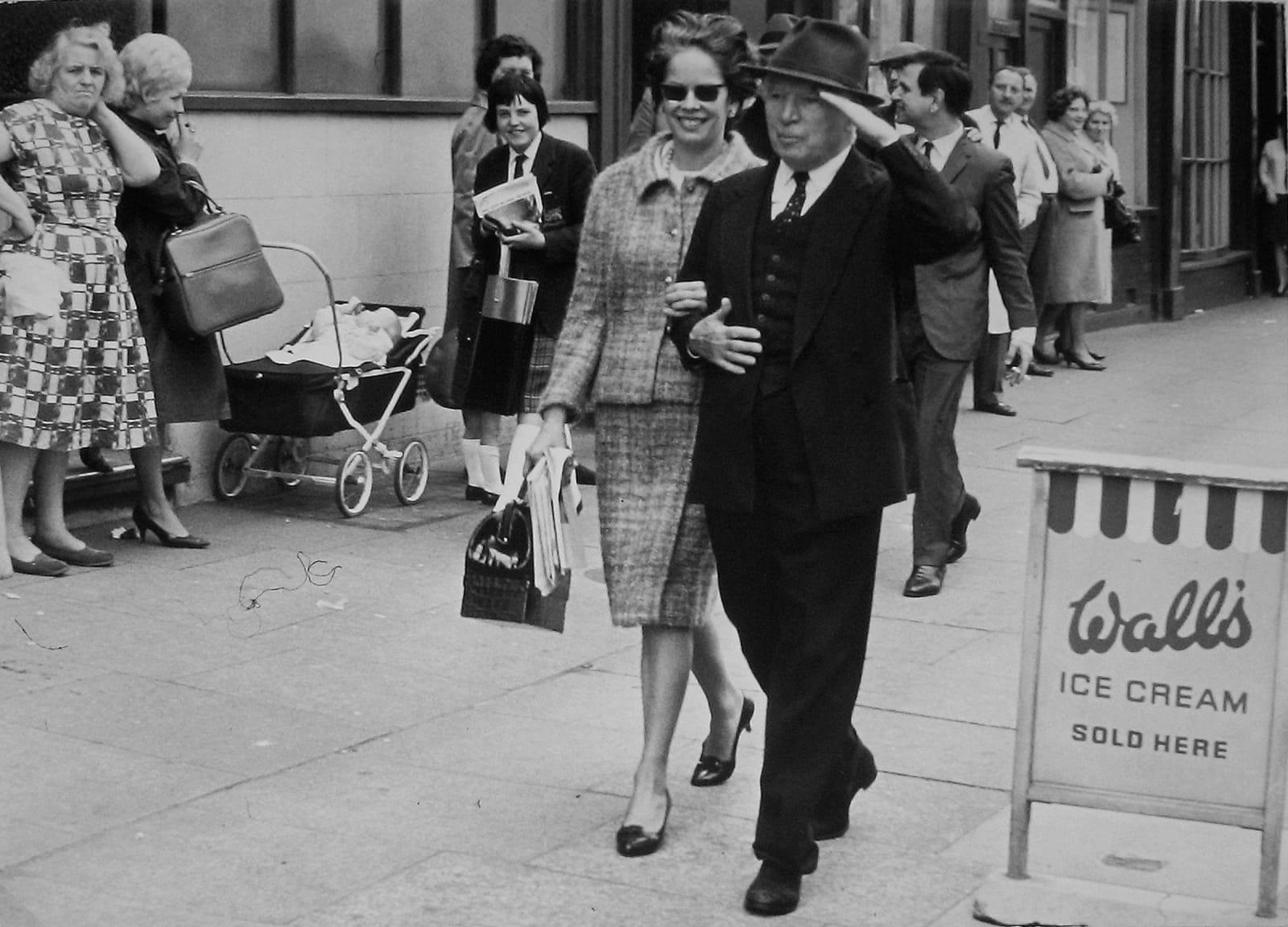 Black and white photograph of Charlie Chaplin walking and linking arms with his wife Oona O’Neill outside at Pinewood Studios, on the set of “A Countess from Hong Kong”