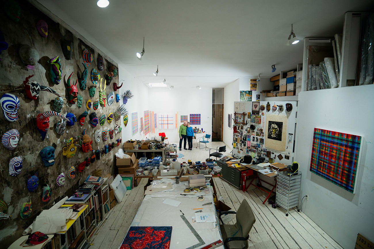 Two artists, Carolyn Oberst and Jeff Way, embracing in a loft space filled with work tables, an array of masks, and paintings