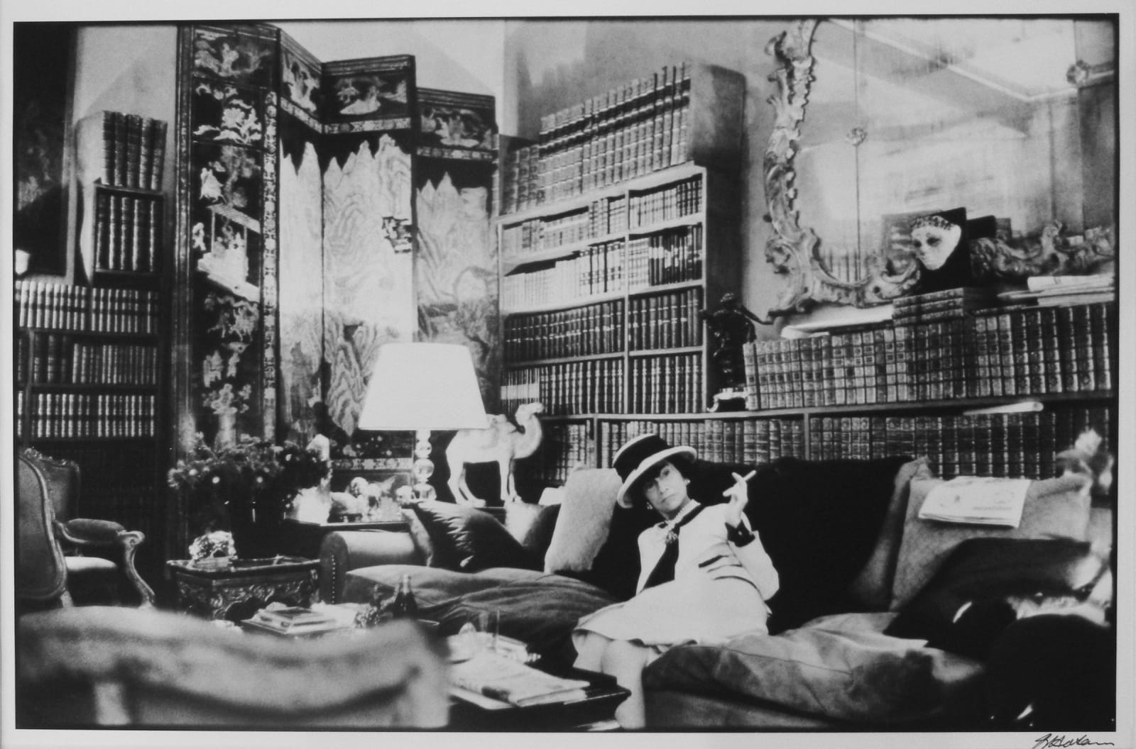 Black and white wide shot photograph of Coco Chanel sitting on the couch and smoking a cigarette in her office at the House of Chanel in Paris