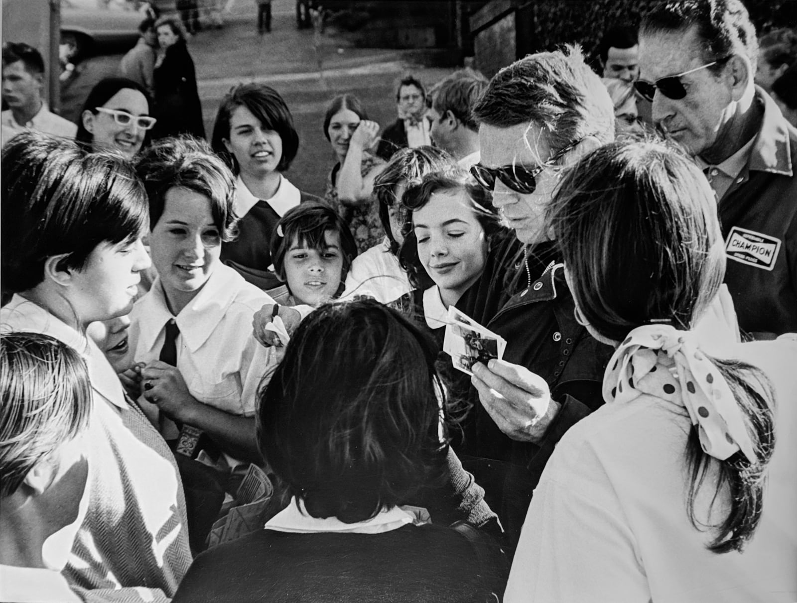 Black and white photograph of Steve Mcqueen signing autographs surrounded by a large group of fans, outside on the set of “Bullitt”