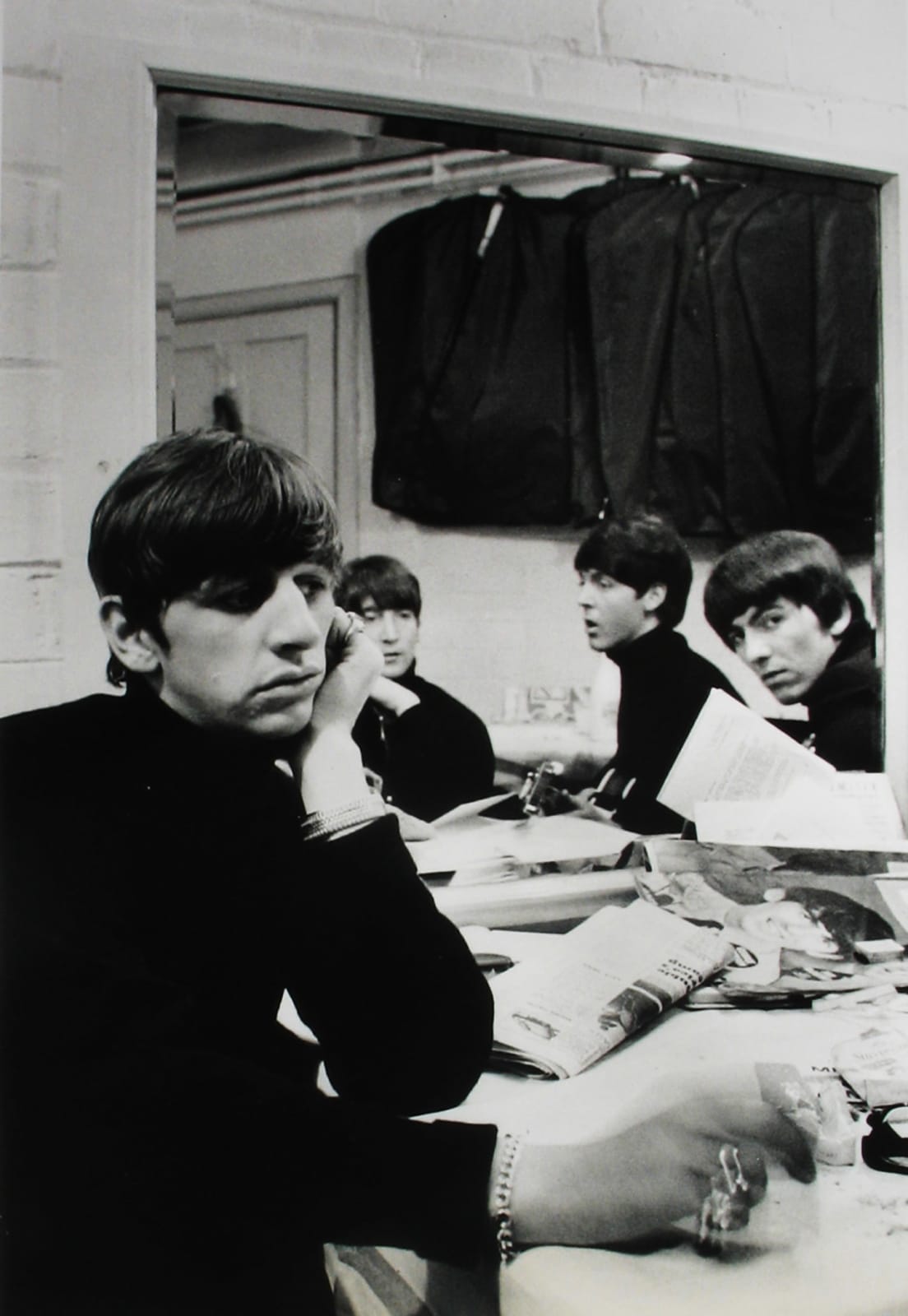 Black and white photograph of The Beatles sitting at a table backstage, all wearing black turtlenecks with Ringo Starr closest to the camera looking out and to the right with his hand on his face.
