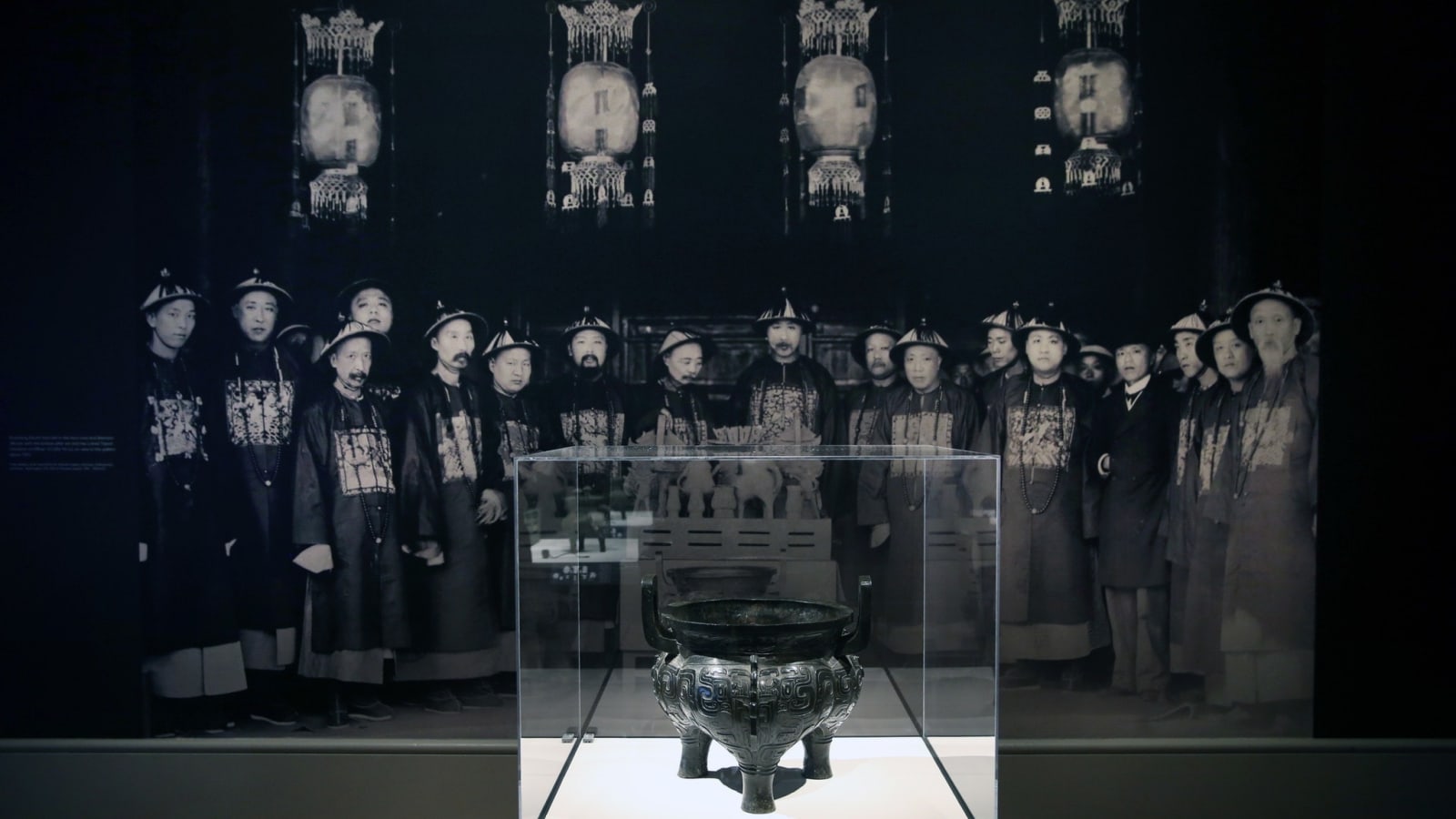 A bronze displayed in the exhibition hall, the background is a group of black and white photos of Qing Dynasty people on the wall