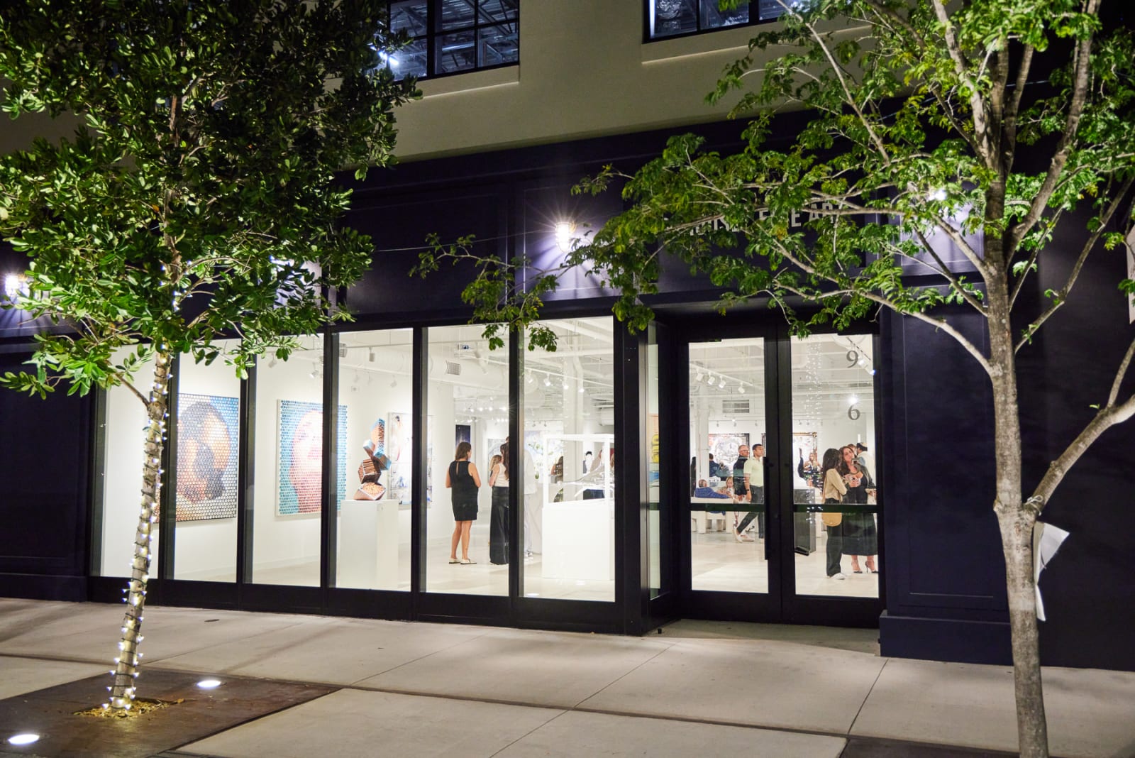 Nighttime photo of NORA District West Palm gallery glowing with Stikki Peaches, Scheltjens, and Jantzen artworks inside.