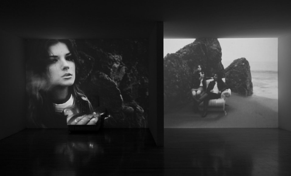 Gallery installation of two screens showing black and white stills. On the left screen, a close up of a face smoking a cigarette. On the right, two figures sit on a chair in front of a boulder.
