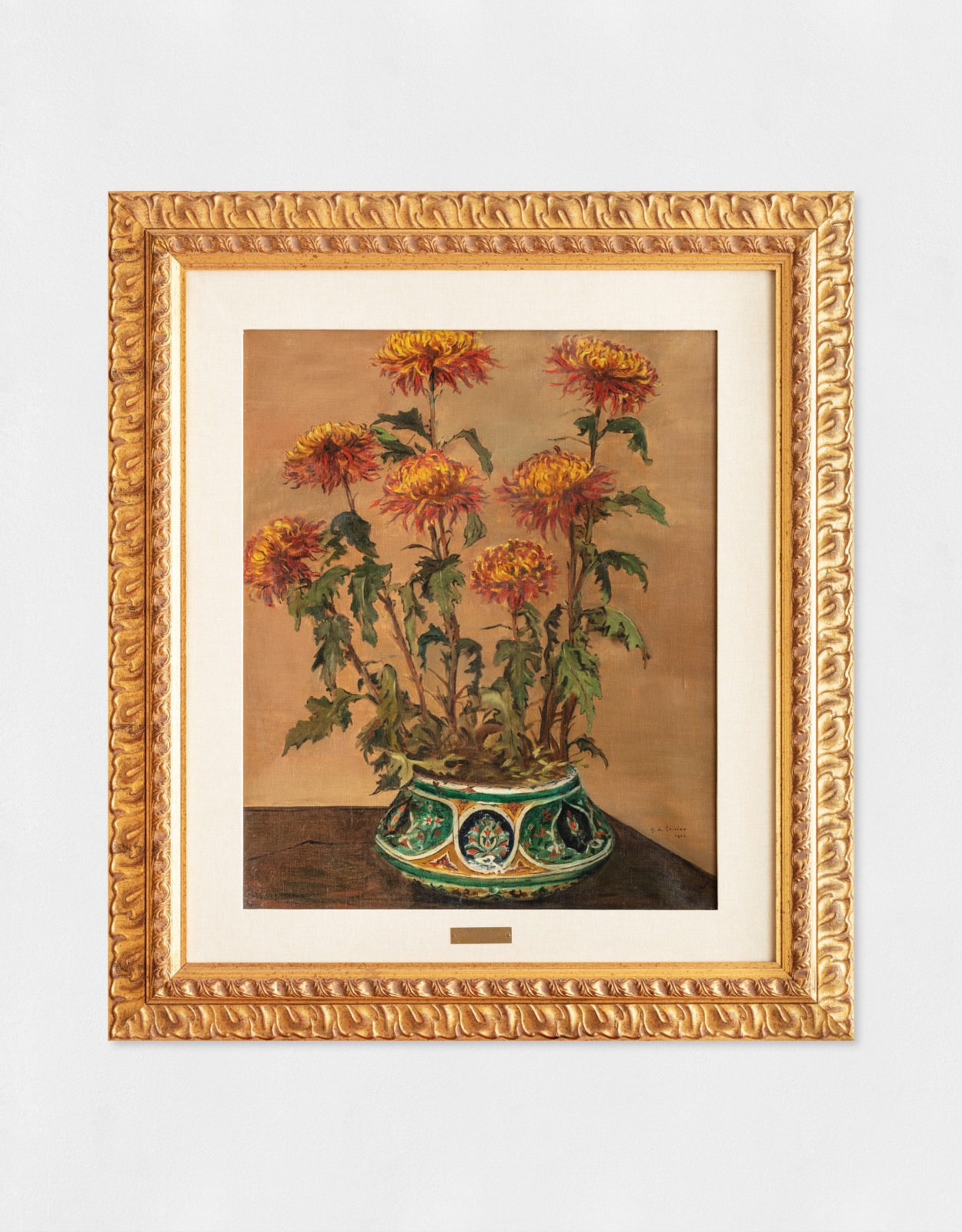 Oil painting of yellow and orange chrysanthemums with green leaves against a beige background in a green and yellow decorative vase positioned on a wooden surface.