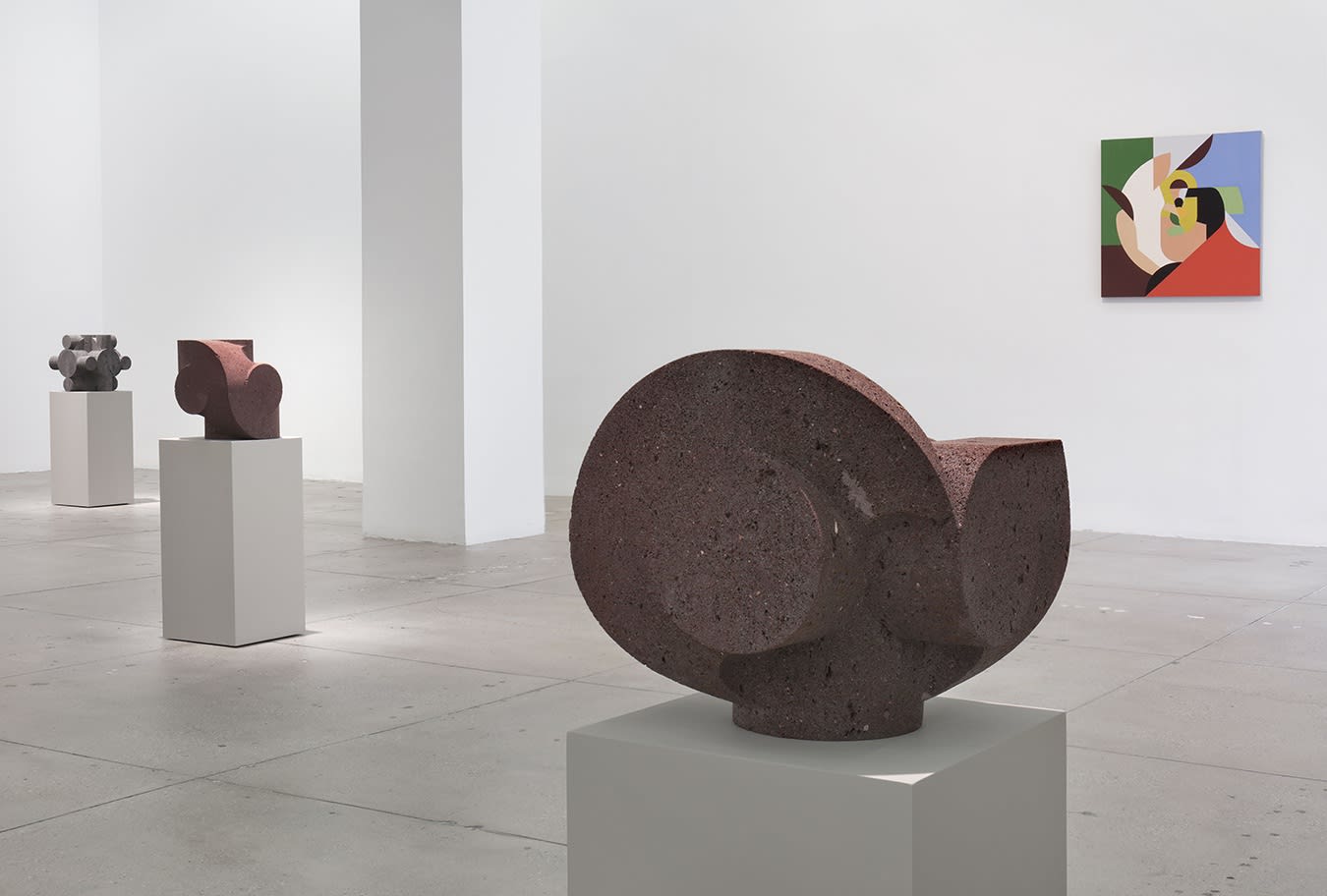 In a large white space, a small abstract painting hangs to the right of 3 small, dark stone sculptures.