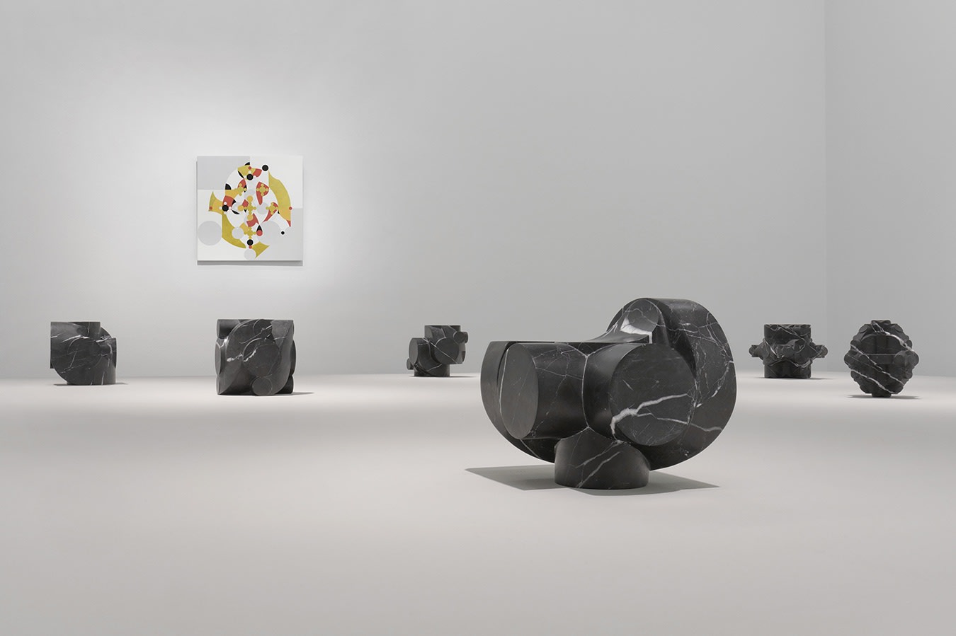 6 small stone sculptures in front of a colorful abstract painting on a white wall.